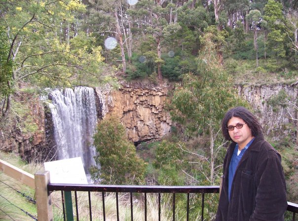 Image of Dr Justin Wejak, he is standing in front of a waterfall 