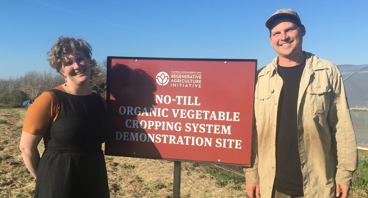 A young woman with short curly hair in an orange tshirt and black jumpsuit and a young man wearing a khaki shirt and cap standing next to a sign saying  "no till organic vegetable cropping system demonstration site" in the background there is a mulched field, a small glimpse of a poly tunnel and a clear blue sky