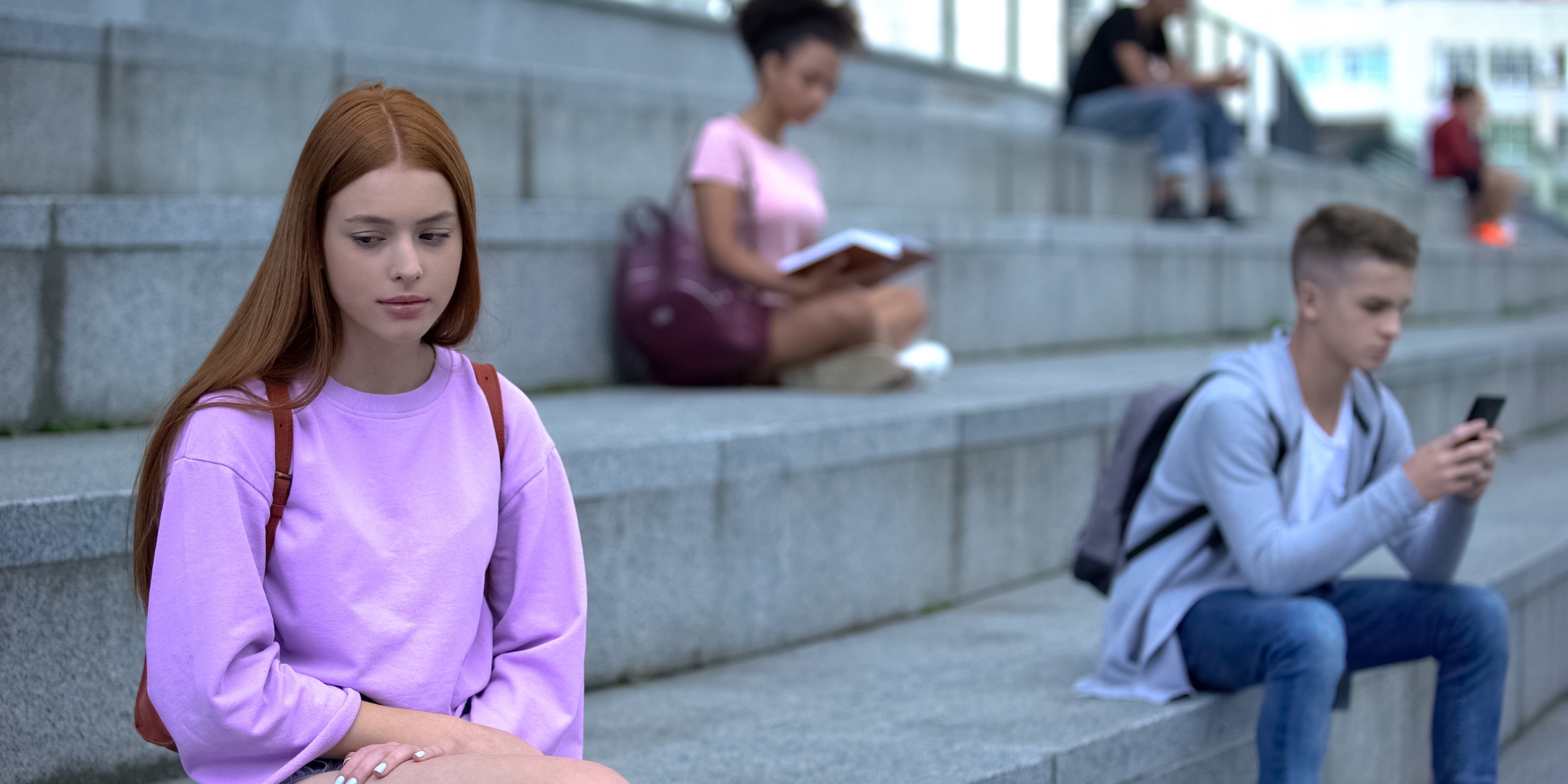 A young women with long red hair sits alone on some grey stone steps, looking downcast. In the background, other young adults sit by themselves, either on their mobile phones or reading.