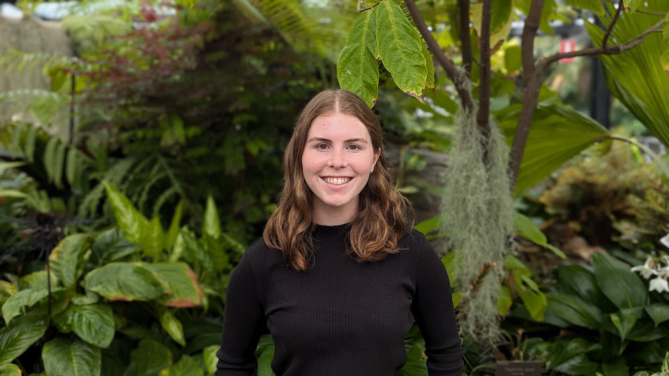 Image of Melissa Ivelja standing in front of a garden of ferns, smiling. 