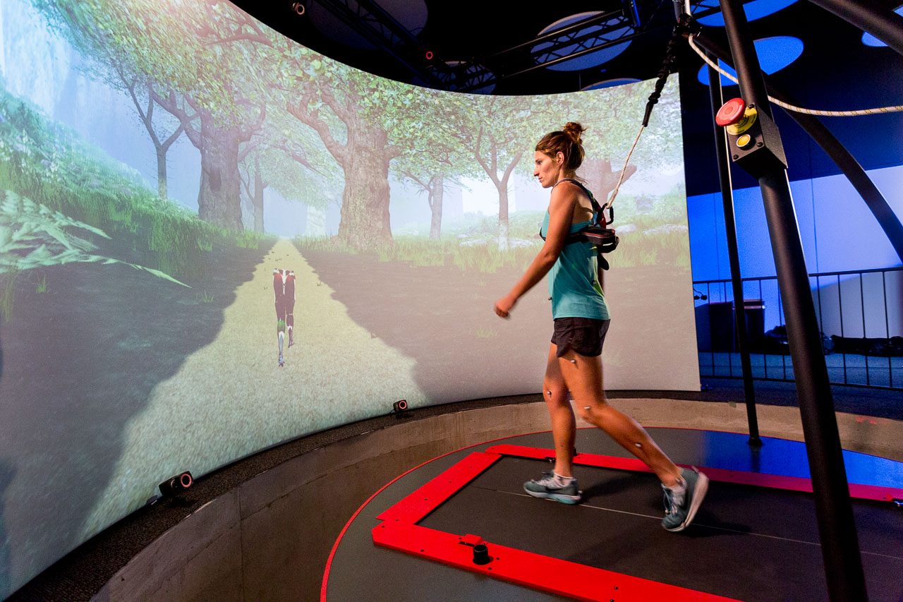 A woman is hooked up to a treadmill-like piece of machinery in front of a large screen displaying a digital image of a forest 