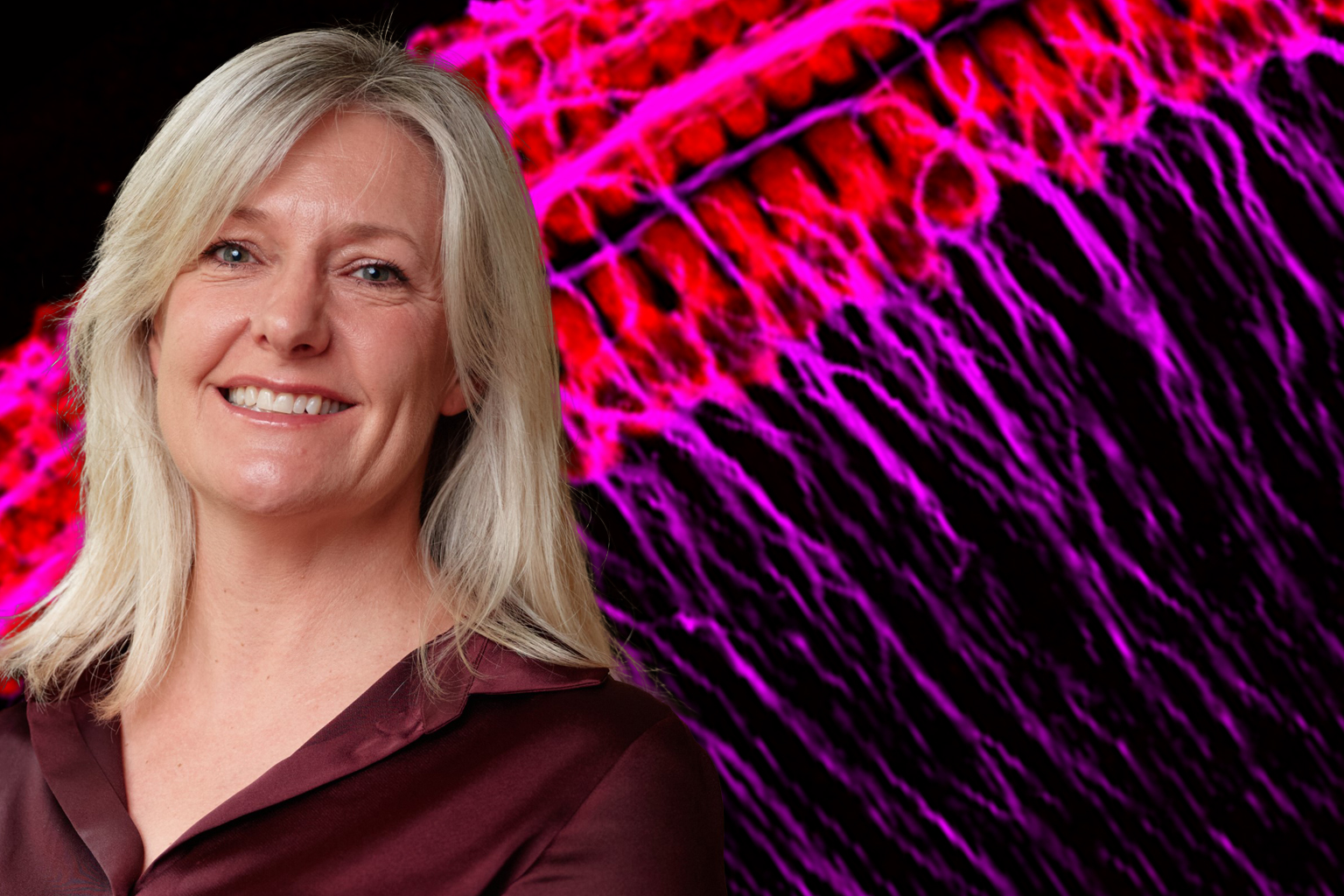 A photo of Bryony, a woman with light hair, smiling at the camera, with a microscopic pink and purple image int he background