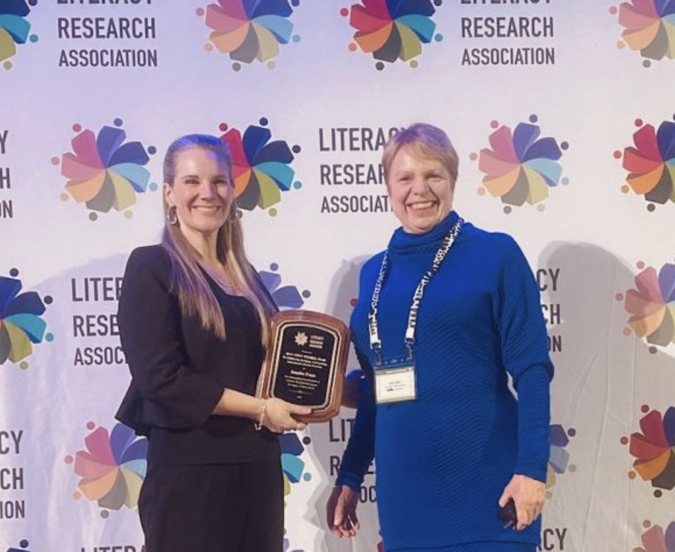 Two women stand on a stage smiling, one holds an award.