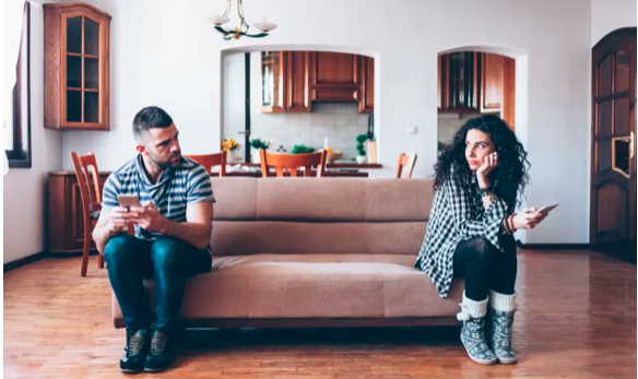 Image of a couple sitting apart at either corners of a sofa looking at each other confrontationally.