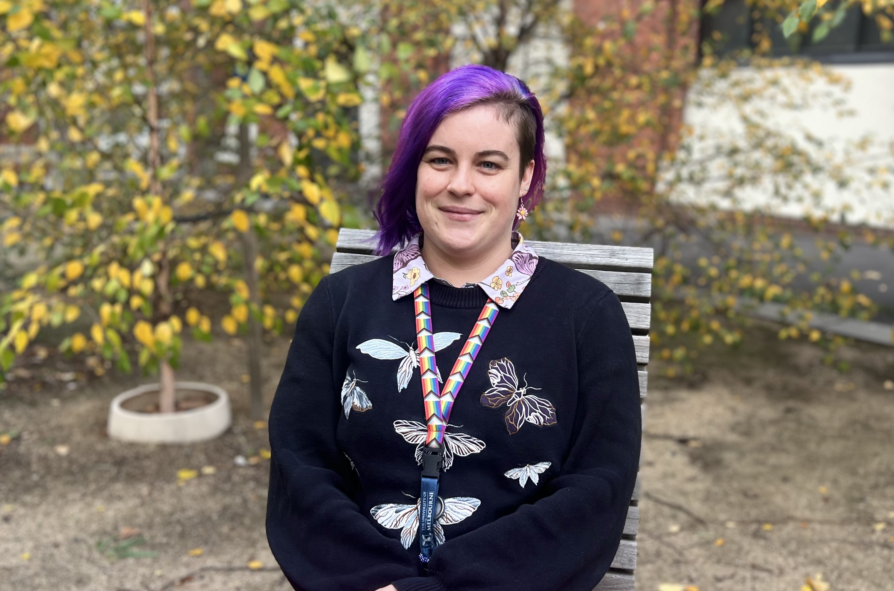 Image of Sarah Timperley, sitting on a chair outside with autumn trees in the background. Sarah is wearing a black top with butterflies on it and has purple hair parted to the left side. 
