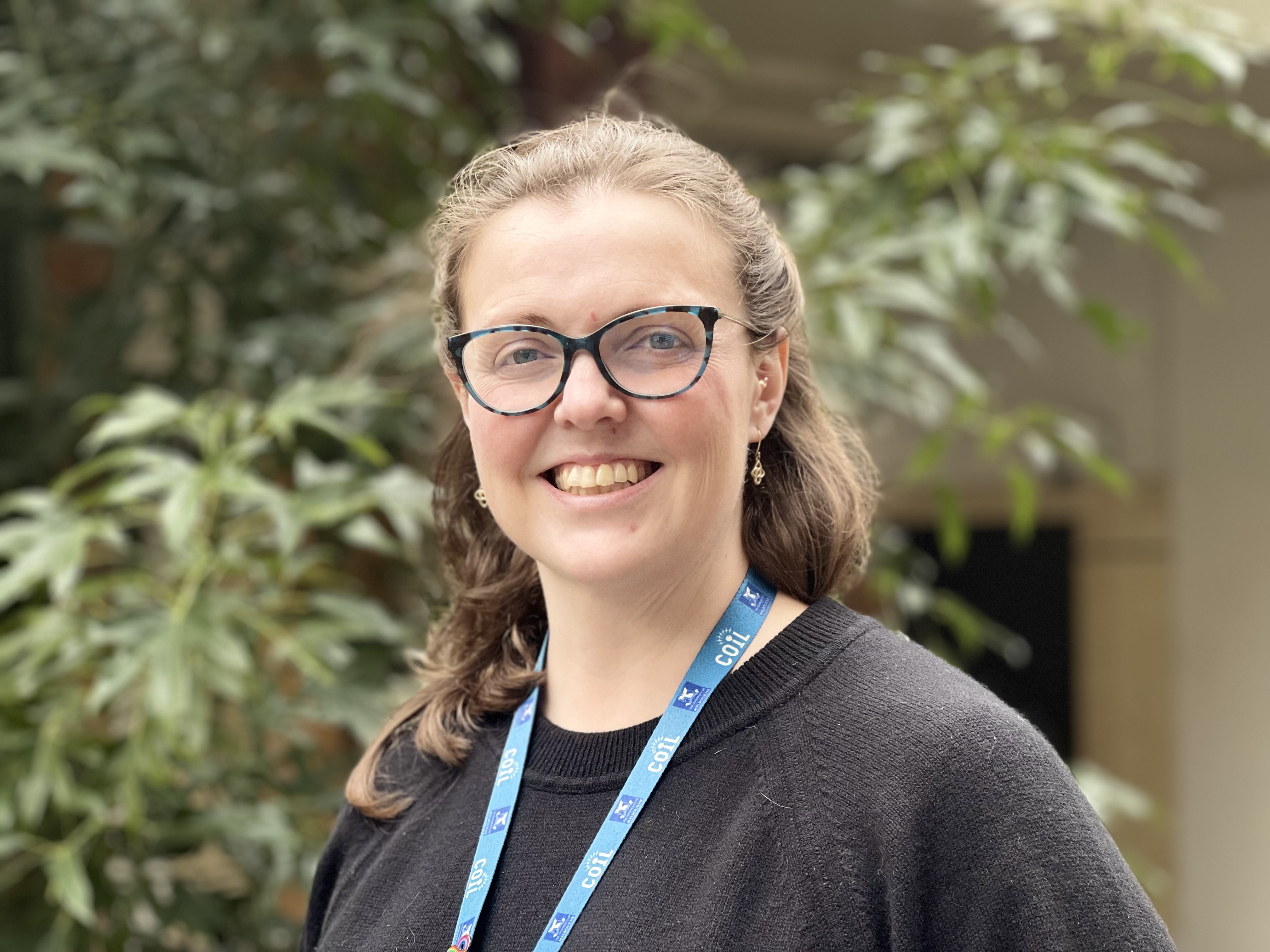 Portrait of Ashley Anderson. She is wearing blue and black speckled glasses and a black top with a blue lanyard. 