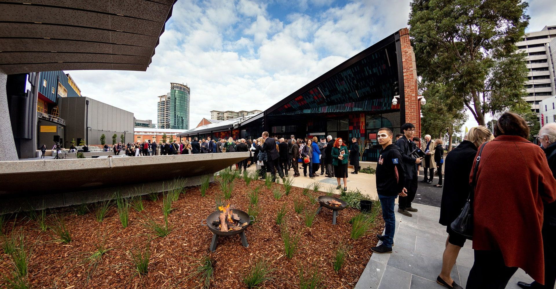 Opening event with crowds outside Faculty of Fine Arts and Music building, with Melbourne city in the background. Fire burning the foreground.
