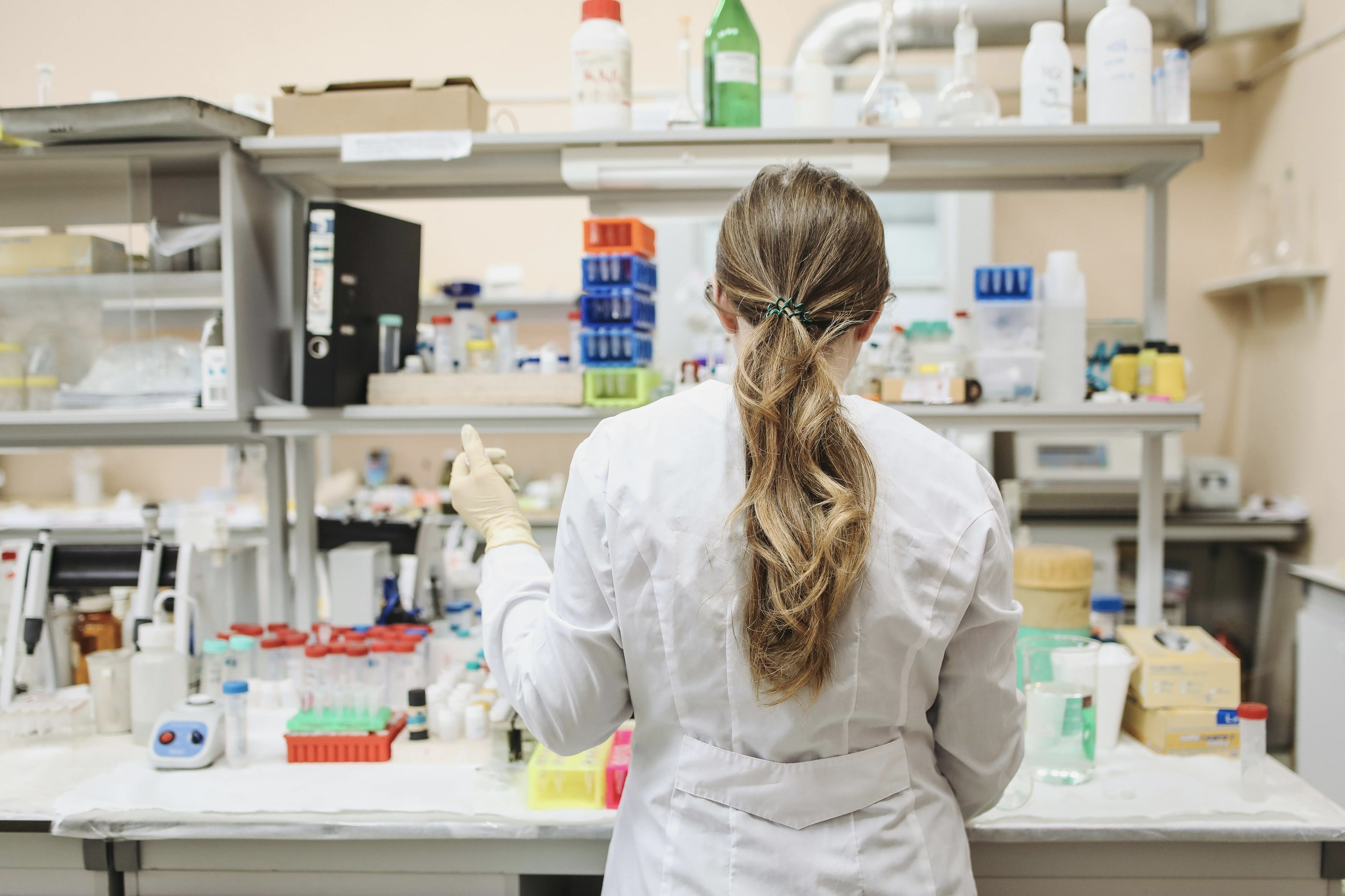 A student wearing a white lab coat using a pipetting device in a research lab. She is facing away from the camera, towards shelving containing bottles of solution and other research equipment.