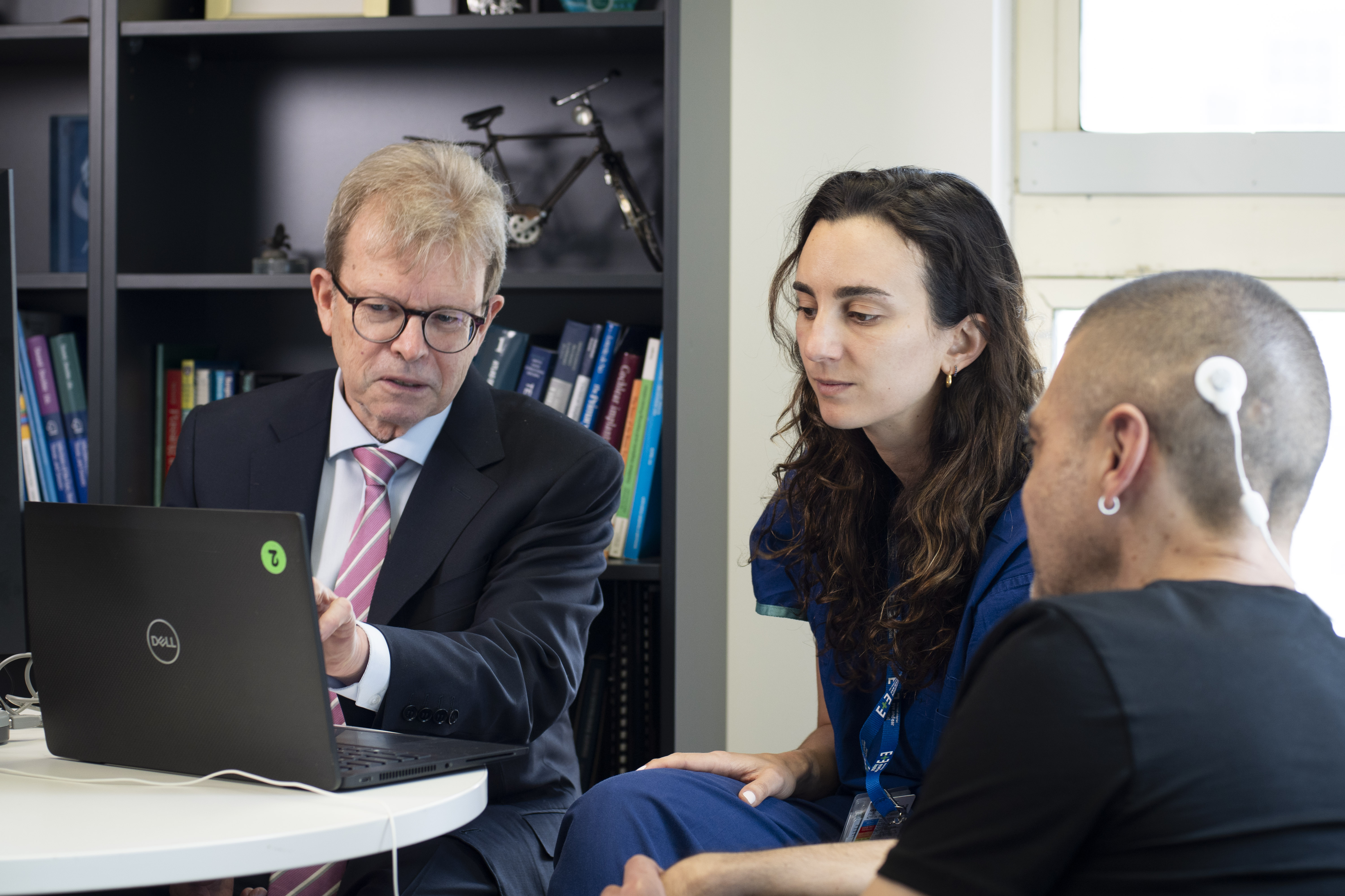 Stephen O'Leary, seated with a nurse and a patient wearing a cochlear implant device, points at a laptop on a table in his office.