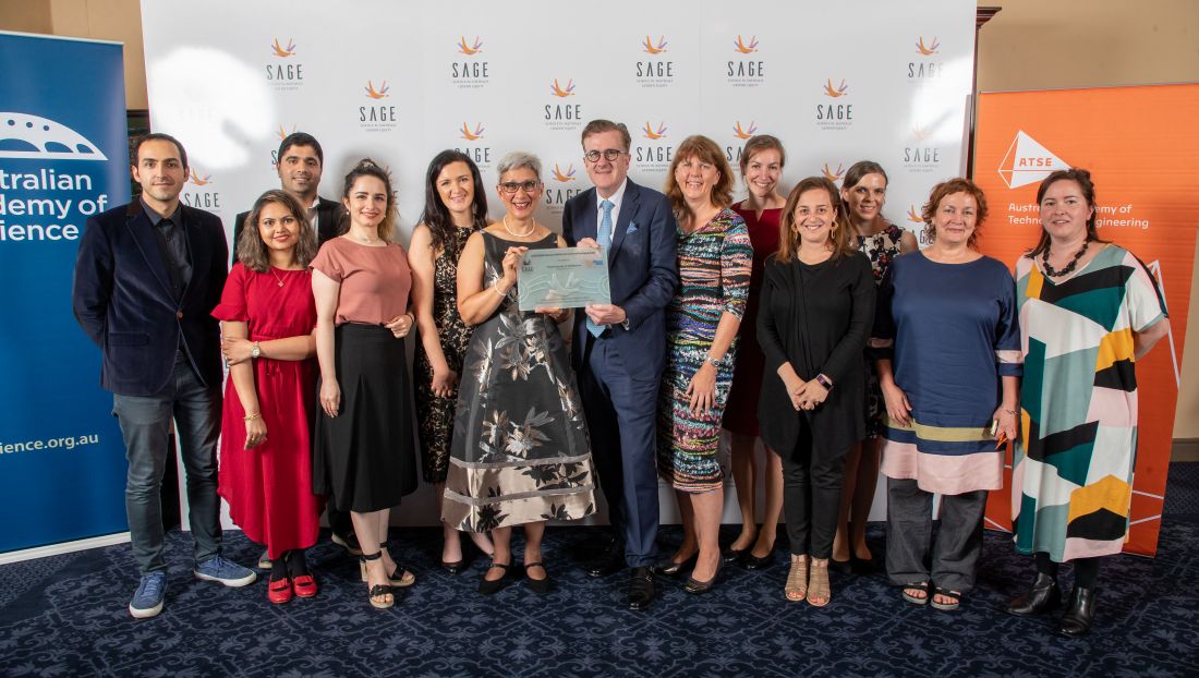 An image from the 2020 Athena SWAN award ceremony. Several members of the Diversity and Inclusion Steering Committee are standing in front of a large white backdrop, emblazoned with the S A G E logo. A man in a suit is presenting Marilys with a glass Athena SWAN Bronze Award. 